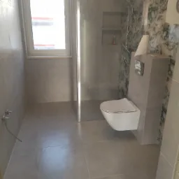 Wykończona łazienka w szarościach z wiszącą toaletą, kabiną prysznicową typu walk-in i ścianą z motywem kwiatowym. Widoczne szare płytki na podłodze i ścianach oraz okno z białymi ramami.