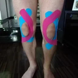 Zbliżenie na nogi mężczyzny z aplikacją kinesiotapingu w kolorach różowym i niebieskim na kolanach, widoczne stopy i fragment podłogi z ciemnego drewna.