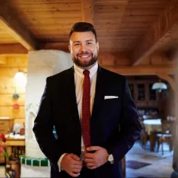 Uśmiechnięty mężczyzna w ciemnym garniturze, białej koszuli i bordowym krawacie w kropki, stojący w rustykalnym wnętrzu z drewnianym sufitem i białym kominkiem.