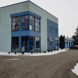 Dwukondygnacyjny budynek w odcieniach błękitu i szarości z dużymi oknami wystawowymi, przed budynkiem zaparkowany samochód, widoczne resztki śniegu.