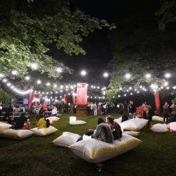 Plenerowa impreza wieczorową porą: ludzie siedzą na podświetlanych pufach na trawie, oświetlona girlandami żarówek, z czerwonymi akcentami dekoracyjnymi w tle.