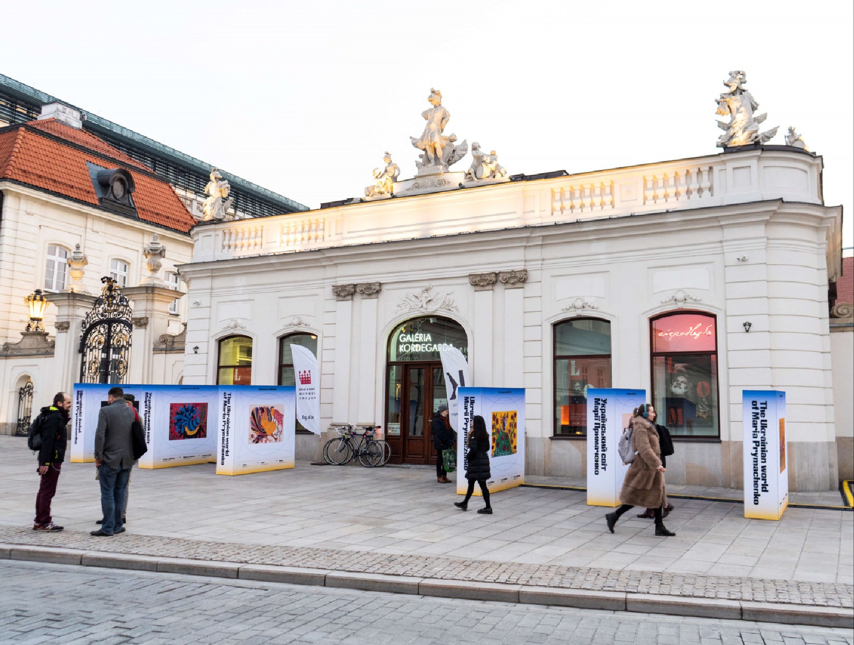 Ekspozycja plenerowa z planszami prezentującymi twórczość Marii Prymachenko przed Galerią Kordegarda w Warszawie, widoczni przechodnie i rower zaparkowany obok.
