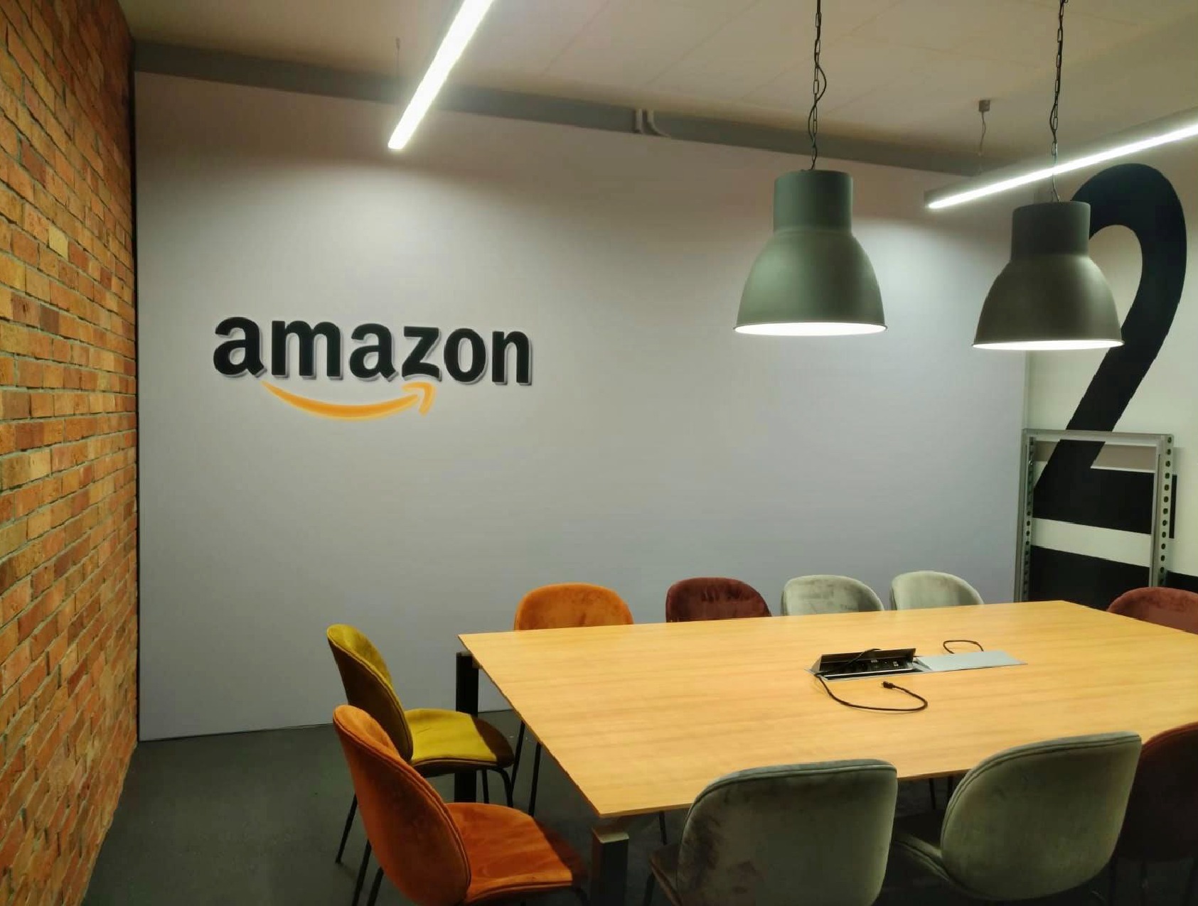 Sala konferencyjna z drewnianym stołem, krzesłami w odcieniach pomarańczu, żółci i szarości, ceglaną ścianą z boku i logo Amazon na ścianie w tle, oświetlona przez wiszące lampy w stylu industrialnym.