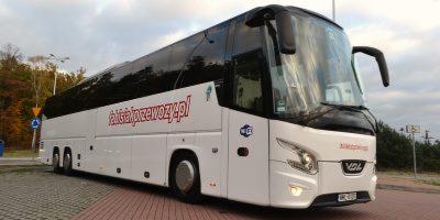 Biały autokar turystyczny z ciemnymi szybami i czerwonym napisem 'faktajakprzewozy.pl' na boku, stojący na parkingu z kostki brukowej, z widocznymi drzewami w tle.