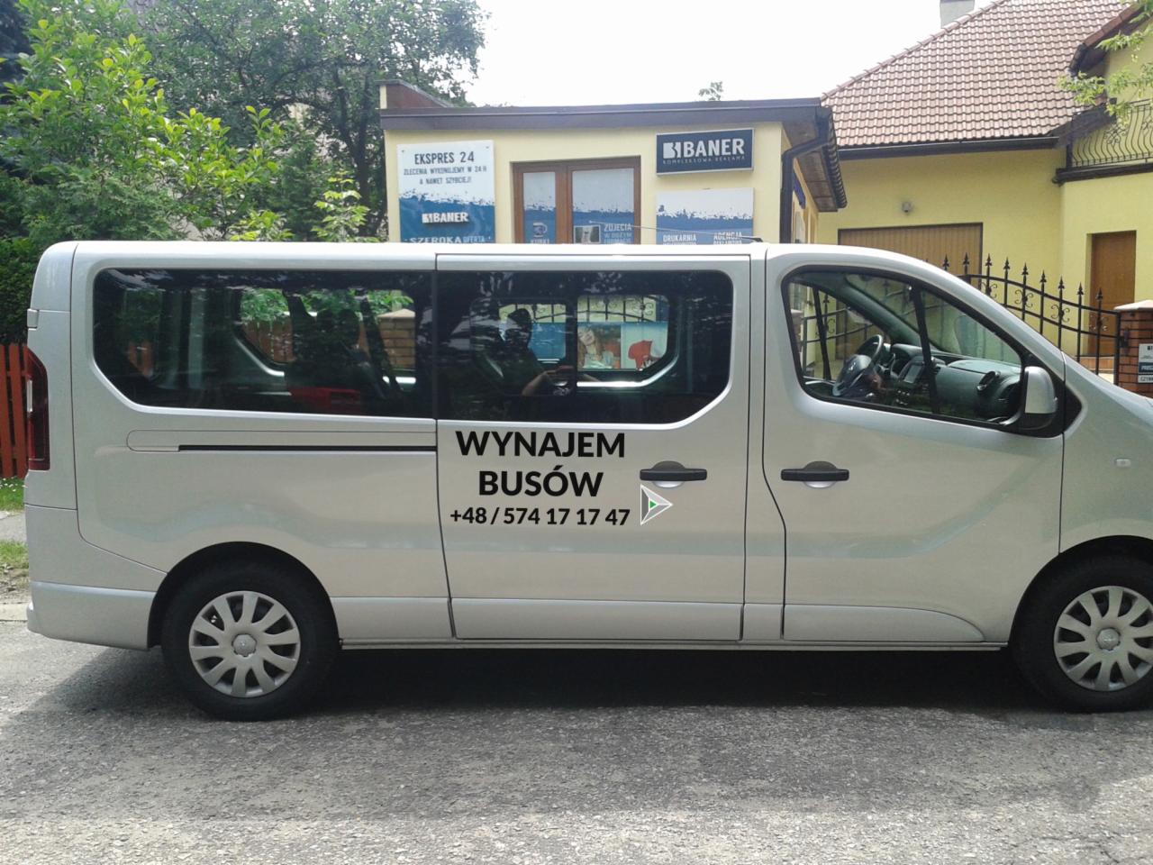 Srebrny bus z napisem 'Wynajem Busów' i numerem telefonu, zaparkowany na ulicy przed budynkiem z szyldami reklamowymi.