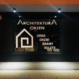 Fasada budynku z czarną stolarką okienną, na której widnieje biały napis 'Architektura Okien' oraz ikona domu, a także informacje o ofercie: okna, drzwi, bramy, rolety.
