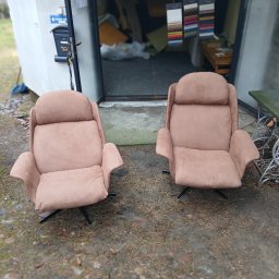 Zakład Tapicerski Feniks - Dwa beżowe fotele z wysokim oparciem na czarnych nogach, stojące przed otwartymi drzwiami warsztatu, w tle wiszące próbniki materiałów i maszyna do szycia przerobiona na stolik.