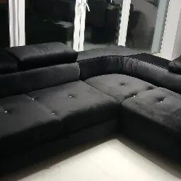 Czarna, welurowa sofa narożna z kryształkami w guziczkach, widoczna w salonie z białymi płytkami podłogowymi i roślinami w tle, na stole narzędzia w skrzynkach.