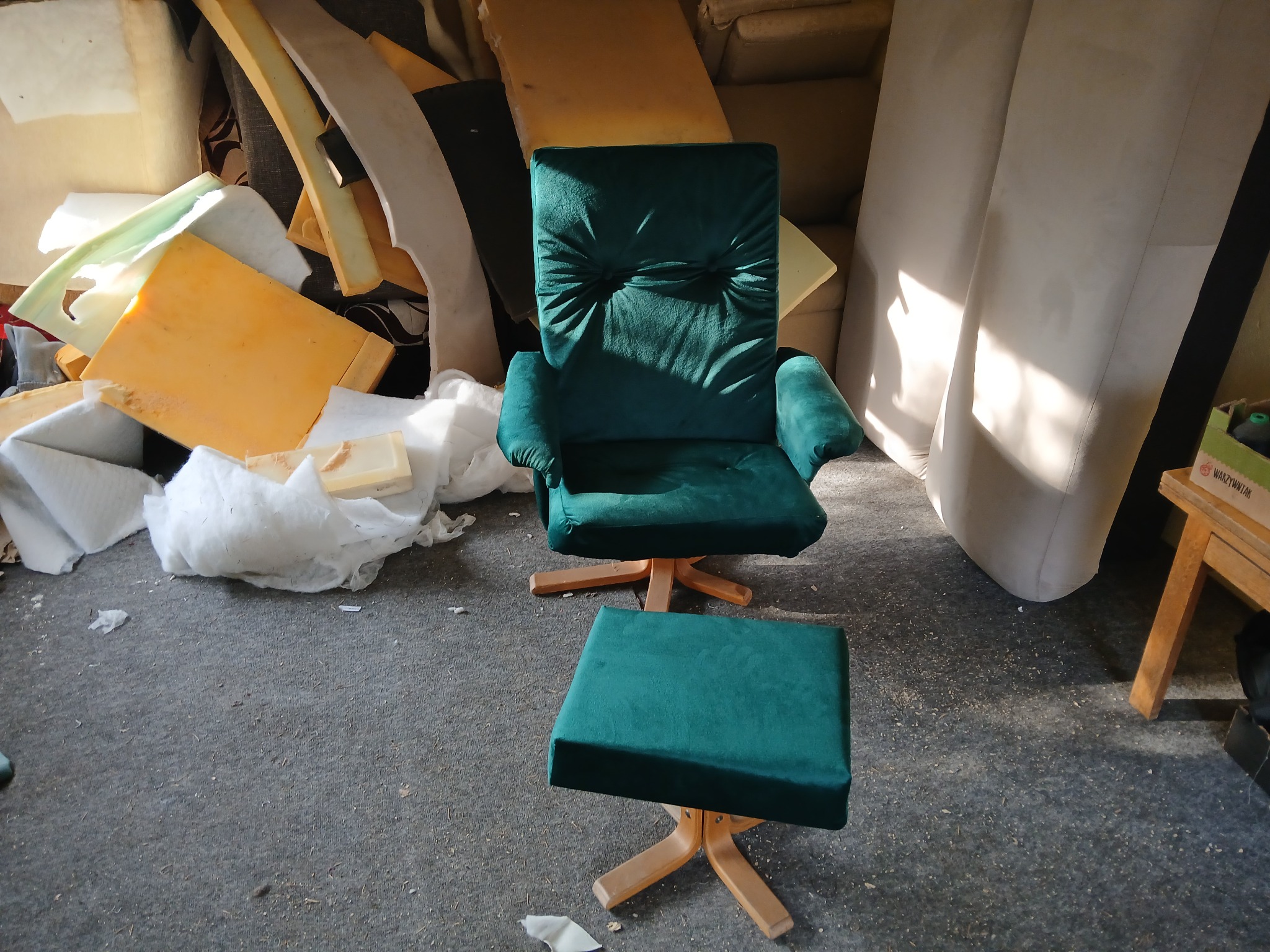 Fotel obrotowy z podnóżkiem w odcieniach szmaragdowej zieleni, ustawiony w pomieszczeniu z widocznymi elementami pianki tapicerskiej w tle, sugerującym warsztat lub magazyn.