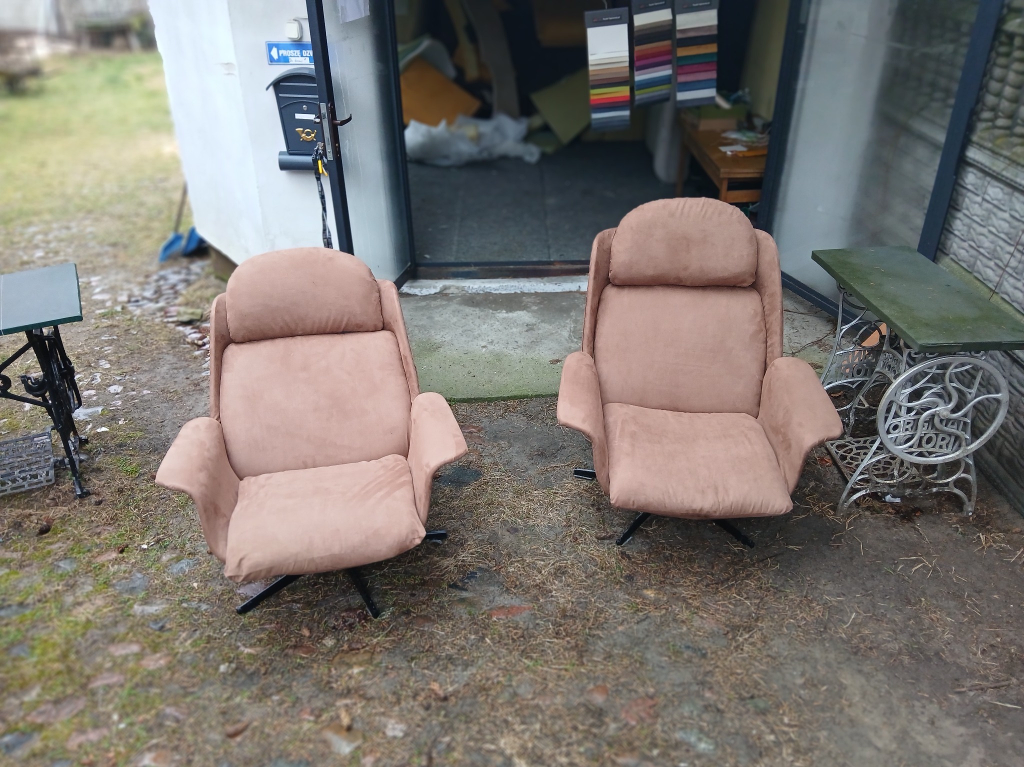 Dwa beżowe fotele z wysokim oparciem na czarnych nogach, stojące przed otwartymi drzwiami warsztatu, w tle wiszące próbniki materiałów i maszyna do szycia przerobiona na stolik.