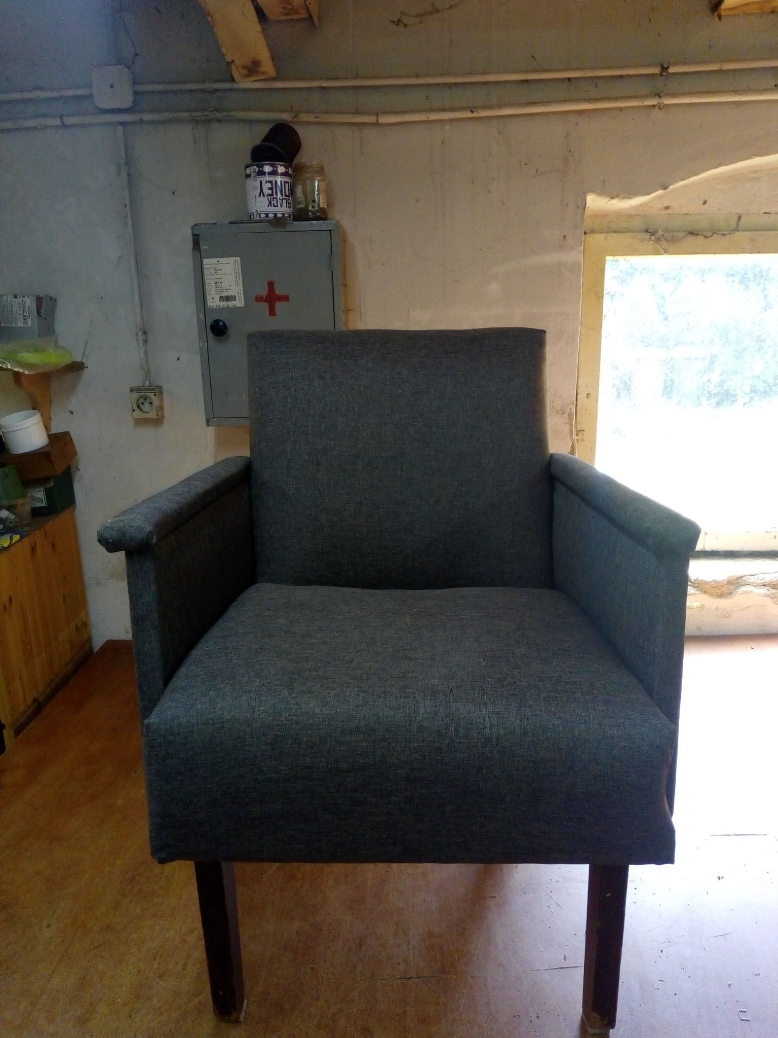 Szary fotel z podłokietnikami stoi na drewnianej podłodze w pomieszczeniu z oknem w tle. Widoczna szafka z apteczką na ścianie.