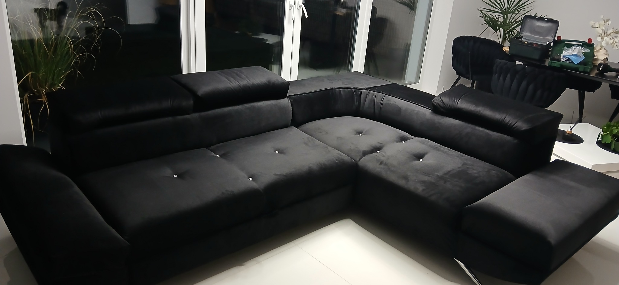 Czarna, welurowa sofa narożna z kryształkami w guziczkach, widoczna w salonie z białymi płytkami podłogowymi i roślinami w tle, na stole narzędzia w skrzynkach.