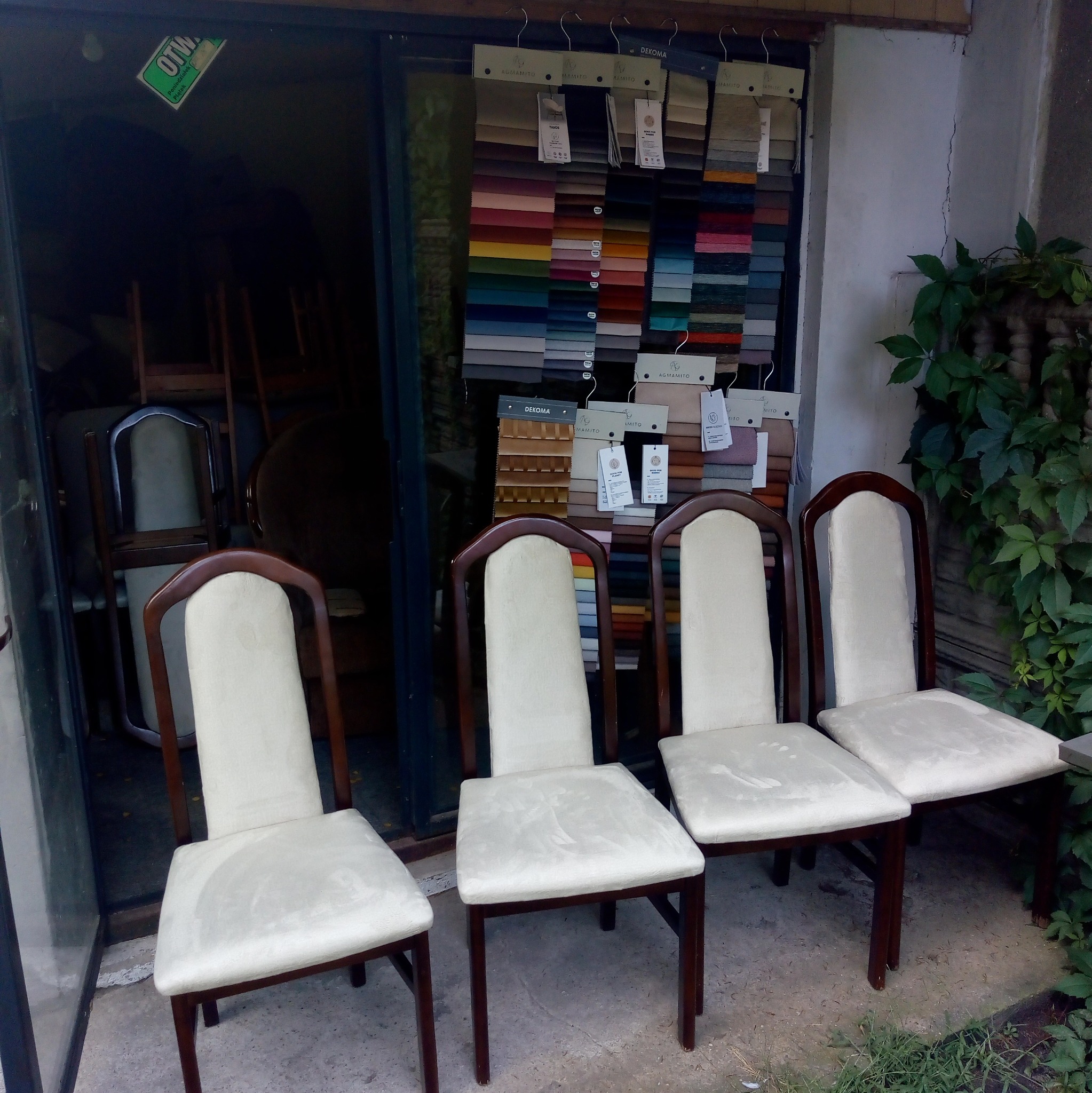 Cztery krzesła z jasną tapicerką ustawione przed sklepem z próbkami tkanin, widoczne ślady użytkowania na siedziskach.