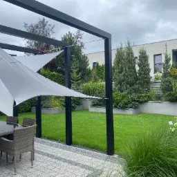 Nowoczesny ogród z równo przystrzyżonym trawnikiem, pergolą z tkaniną zacieniającą i meblami ogrodowymi. W tle budynek mieszkalny i różnorodne nasadzenia.