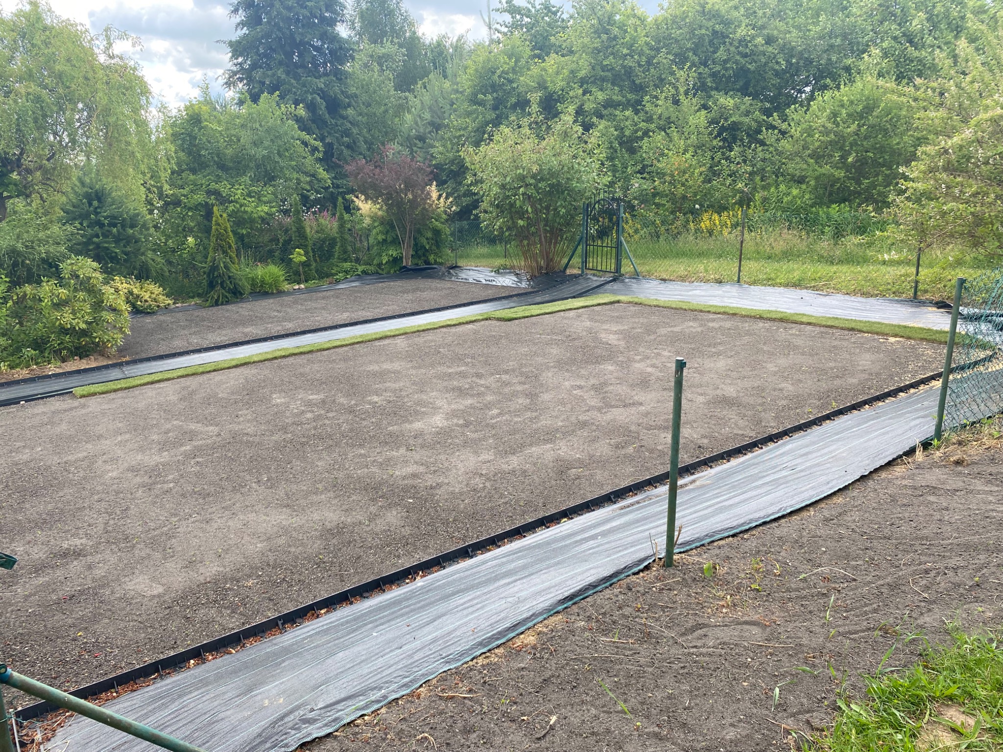 Przygotowanie terenu pod trawnik z rolki: rozłożona agrowłóknina i obrzeża. Widoczne fragmenty trawy w rolkach, zielona brama w tle i drzewa.