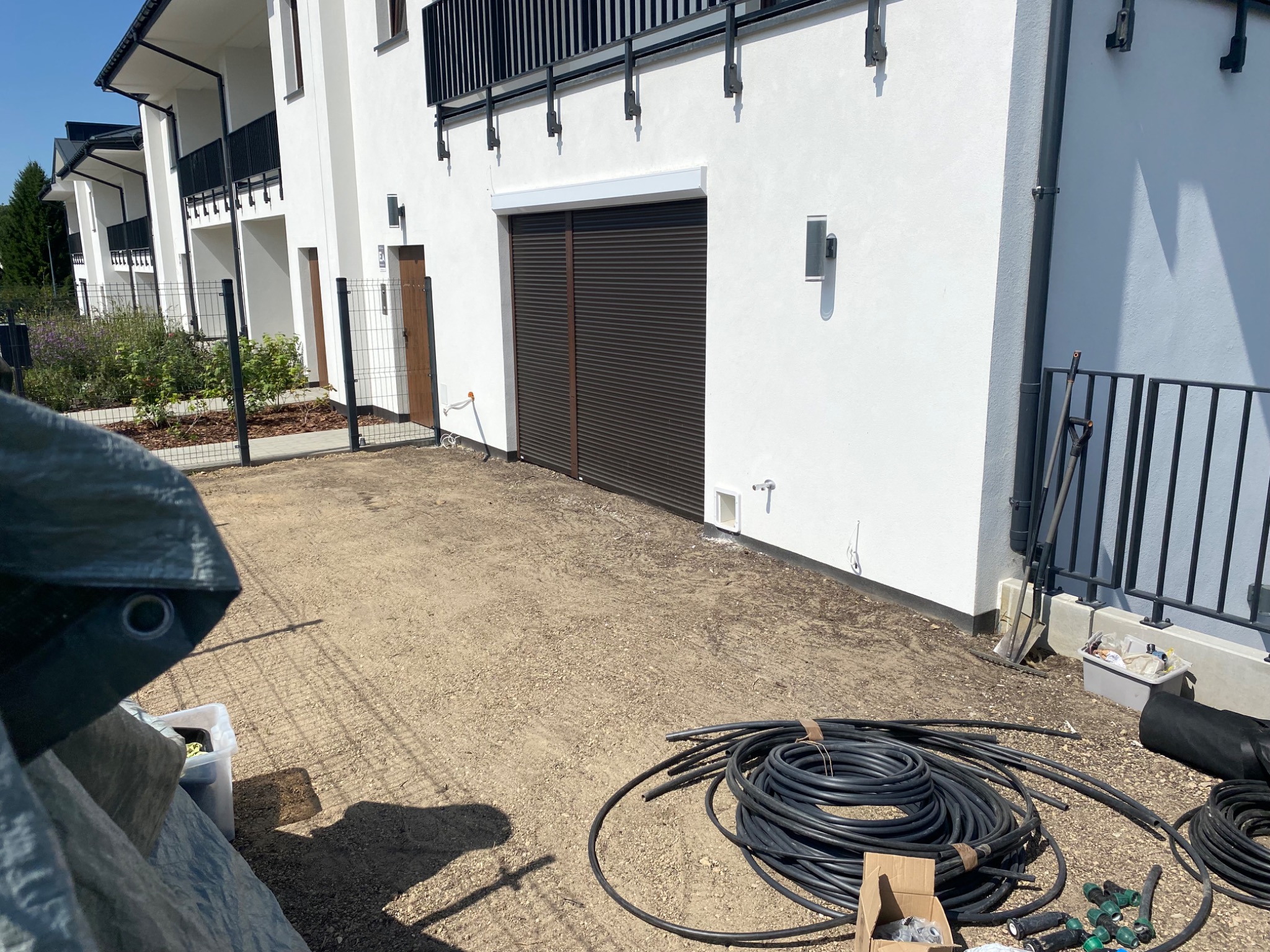 Przygotowania do instalacji nawadniania: rury, złączki na żwirowym podłożu przed nowoczesnym budynkiem z brązową bramą rolowaną i metalowym ogrodzeniem.