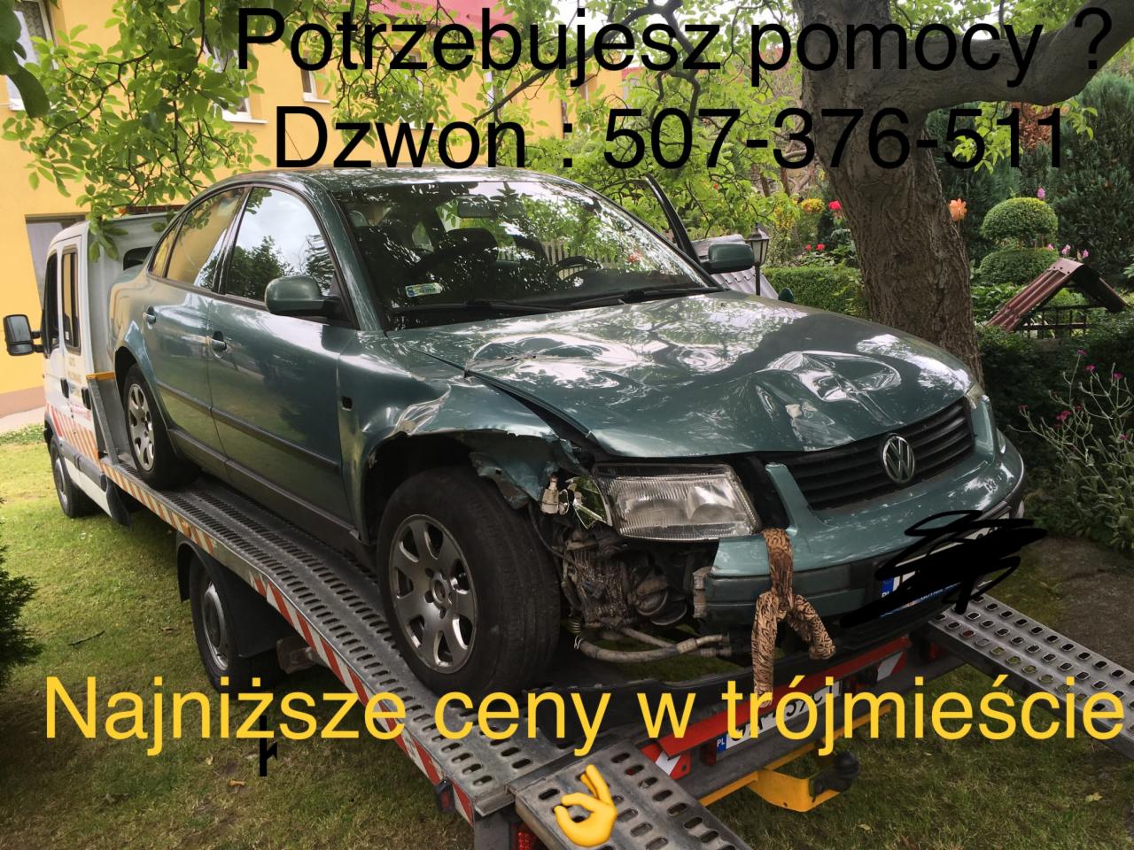 Uszkodzony zielony Volkswagen Passat na platformie białej lawety, z widocznym numerem telefonu i hasłem 'Najniższe ceny w Trójmieście' na tle ogrodu.