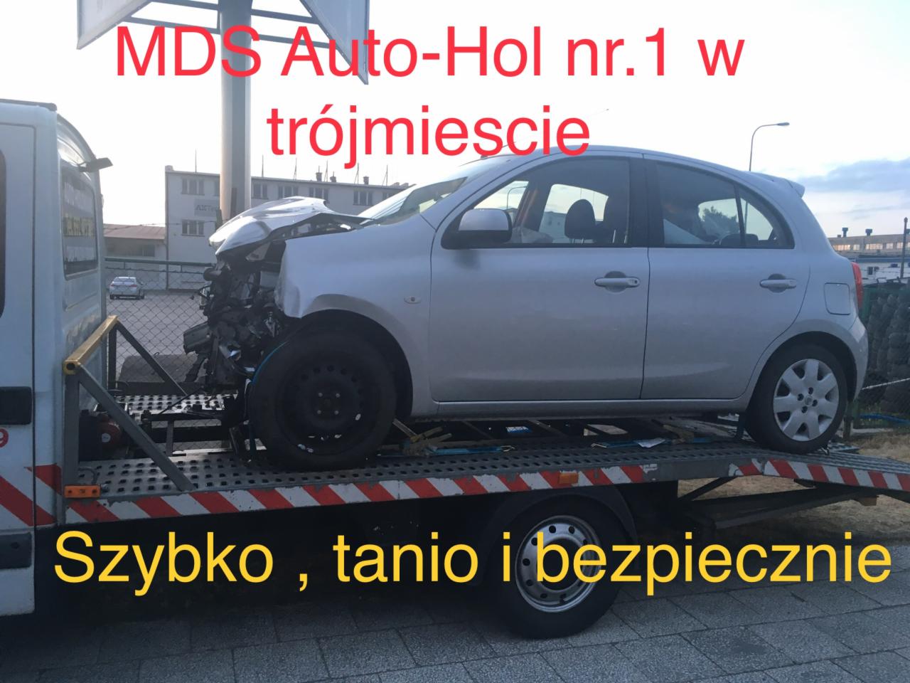 Srebrny samochód osobowy z poważnymi uszkodzeniami przodu, załadowany na platformę lawety z czerwono-białymi pasami, widoczny napis reklamowy 'MDS Auto-Hol nr.1 w trójmieście' oraz hasło 'Szybko...