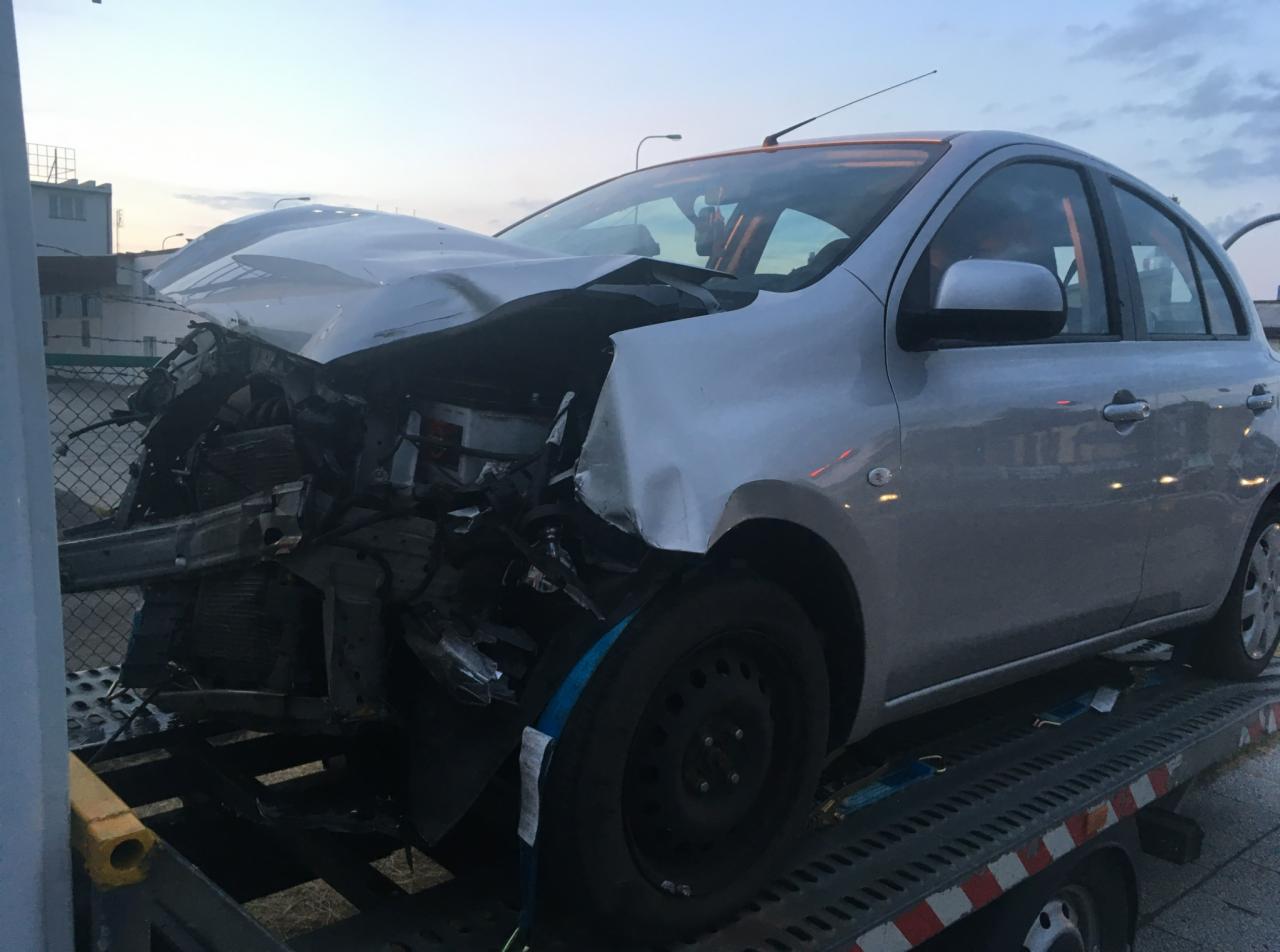 Srebrny samochód osobowy z poważnymi uszkodzeniami przodu, załadowany na platformę lawety. Widoczne zgniecione elementy karoserii i uszkodzone podzespoły. Ujęcie z boku, o zmierzchu.