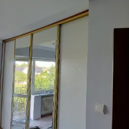 Nowoczesna szafa wnękowa z przesuwnymi drzwiami lustrzanymi i złotymi ramami, widoczna w trakcie prac wykończeniowych, zabezpieczona folią podłoga.