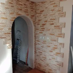 Wykończenie ściany imitacją kamienia wok&oacute;ł łukowego przejścia, widoczna drabina w tle i przełączniki światła na ścianie.