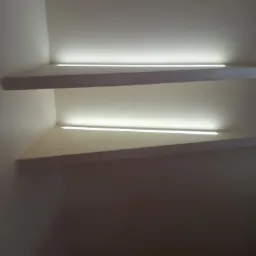Dwie białe półki narożne z ukrytym oświetleniem LED w rogu pokoju, widok z dołu, ciepła barwa światła.