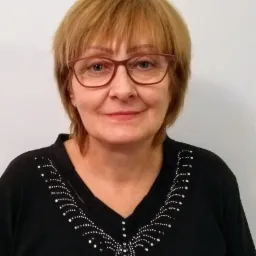 Portret kobiety w okularach z krótkimi blond włosami, uśmiechniętej, ubranej w czarną bluzkę z ozdobnym wzorem z białych kropek.