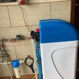 Domowa stacja uzdatniania wody z niebiesko-białą obudową, widoczne rury, filtry, zawory i narzędzie do montażu filtrów, na tle płytek ceramicznych i wyłącznika prądu.