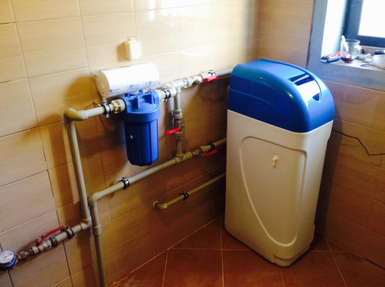 System filtracji wody z niebieskim filtrem i białym zmiękczaczem w rogu pomieszczenia z brązowymi płytkami, widoczne rury i zawory.