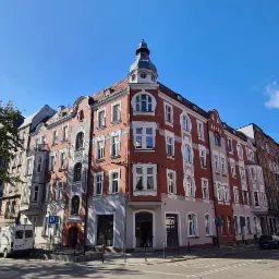 Kamienica narożna z wieżyczką w stylu historyzmu, odnowiona elewacja z czerwonej cegły i białymi detalami, błękitne niebo z chmurami w tle.