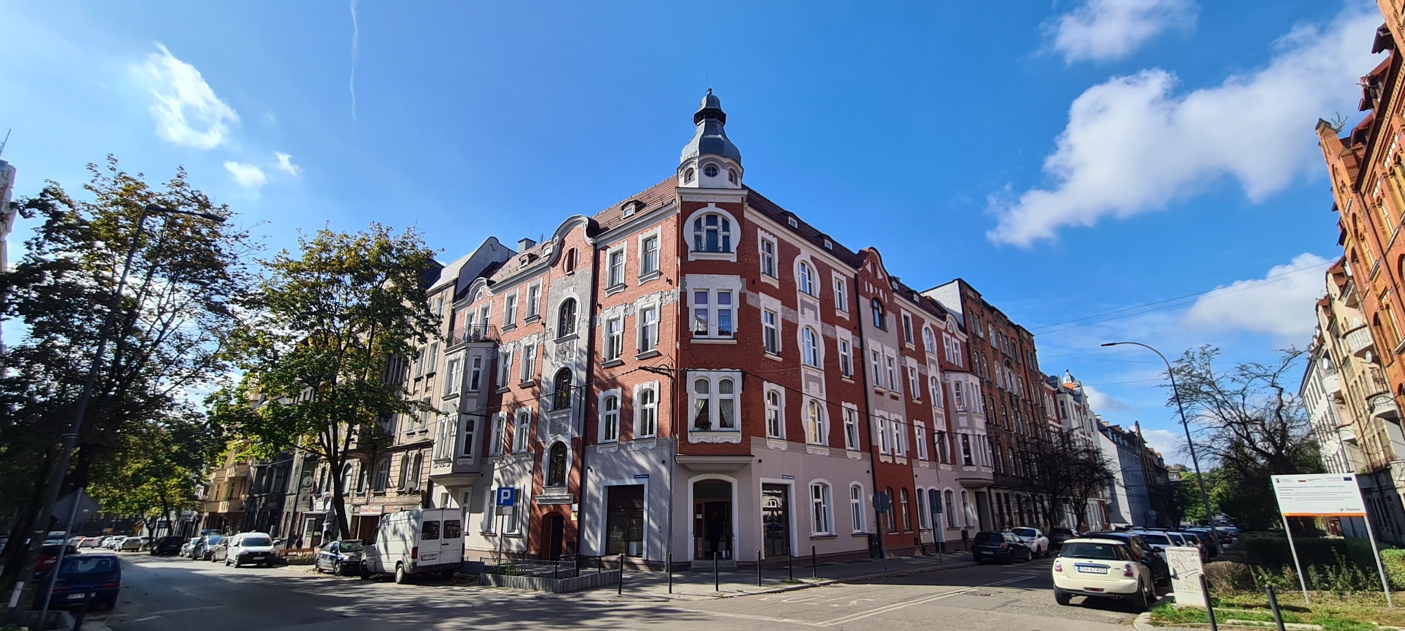 Kamienica narożna z wieżyczką w stylu historyzmu, odnowiona elewacja z czerwonej cegły i białymi detalami, błękitne niebo z chmurami w tle.