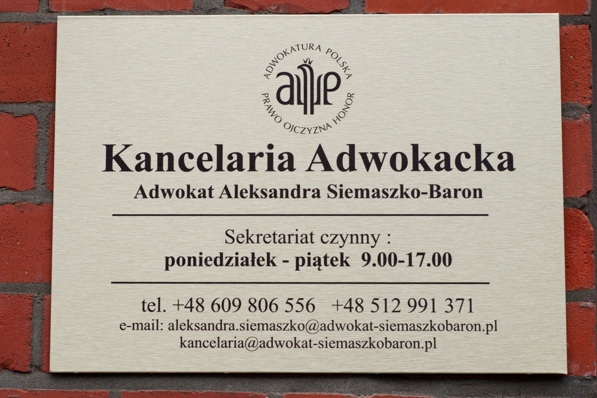 Tabliczka informacyjna na ceglanej ścianie z napisem 'Kancelaria Adwokacka Adwokat Aleksandra Siemaszko-Baron', logo Adwokatury Polskiej, dane kontaktowe i godziny pracy sekretariatu.