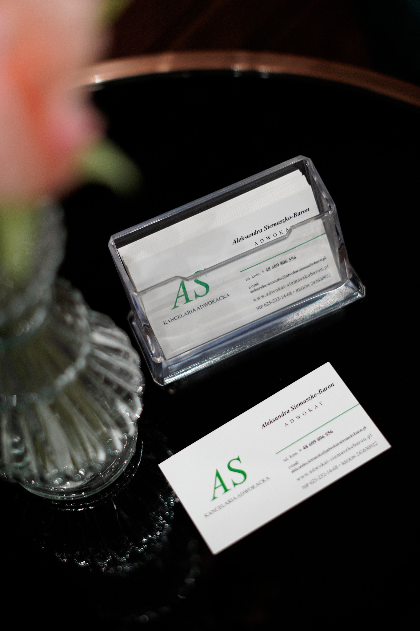 Wizytówki adwokackie w przezroczystym podajniku i pojedyncza wizytówka leżąca obok, na błyszczącym czarnym blacie, obok dekoracyjny szklany wazon.