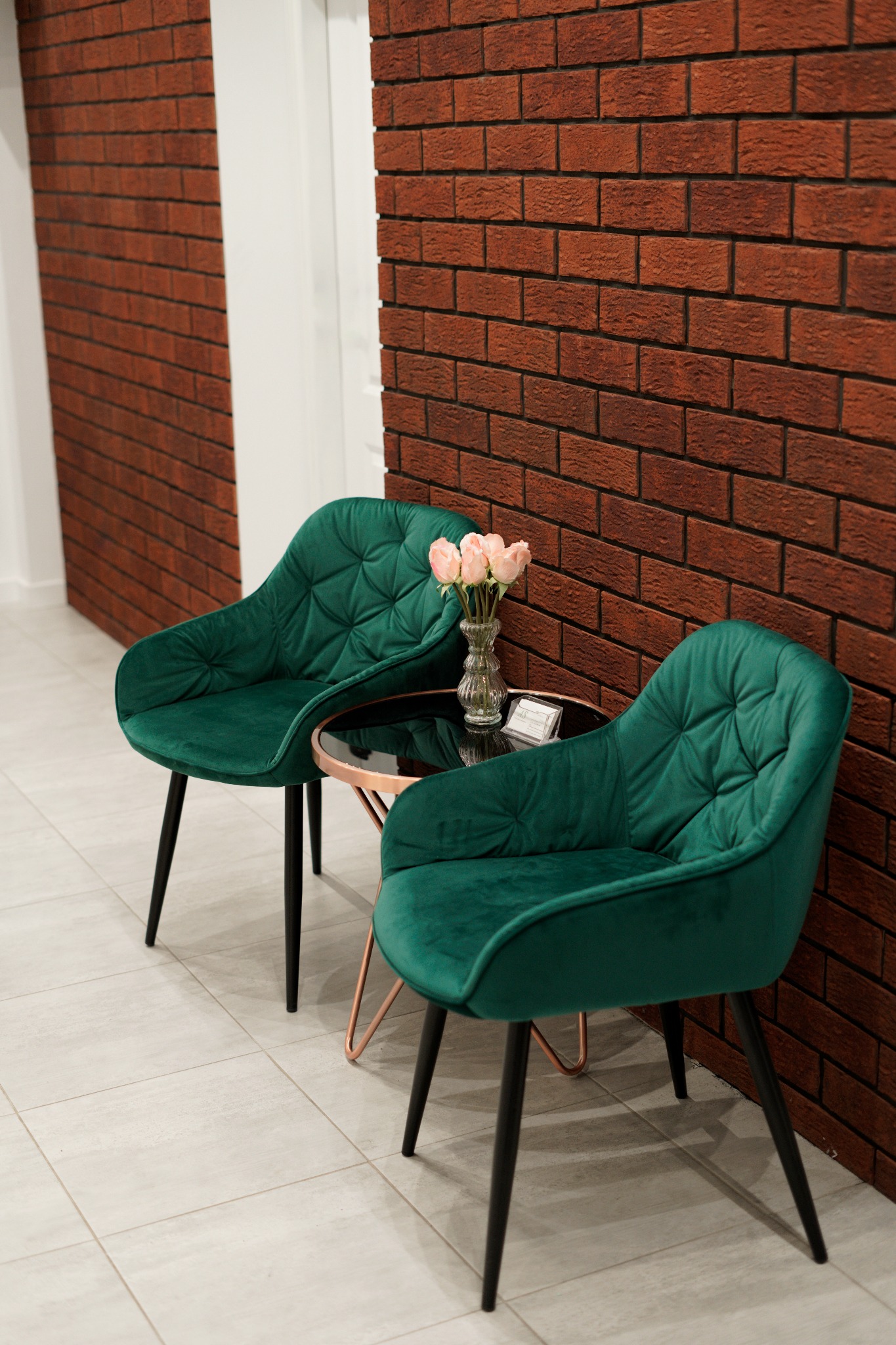 Wnętrze kancelarii: Dwa zielone, welurowe fotele z pikowaniem, stolik kawowy z czarnym blatem i miedzianą podstawą, wazon z różowymi różami, ceglana ściana w tle.