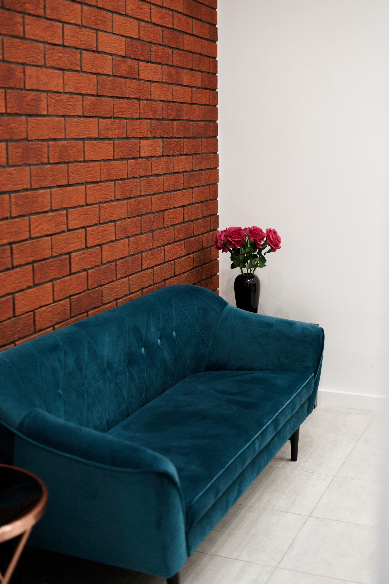 Turkusowa, welurowa sofa z guzikami na tle ceglanej ściany i białej ściany, z czarnym wazonem z różowymi różami, ustawiona na jasnej podłodze z płytek.