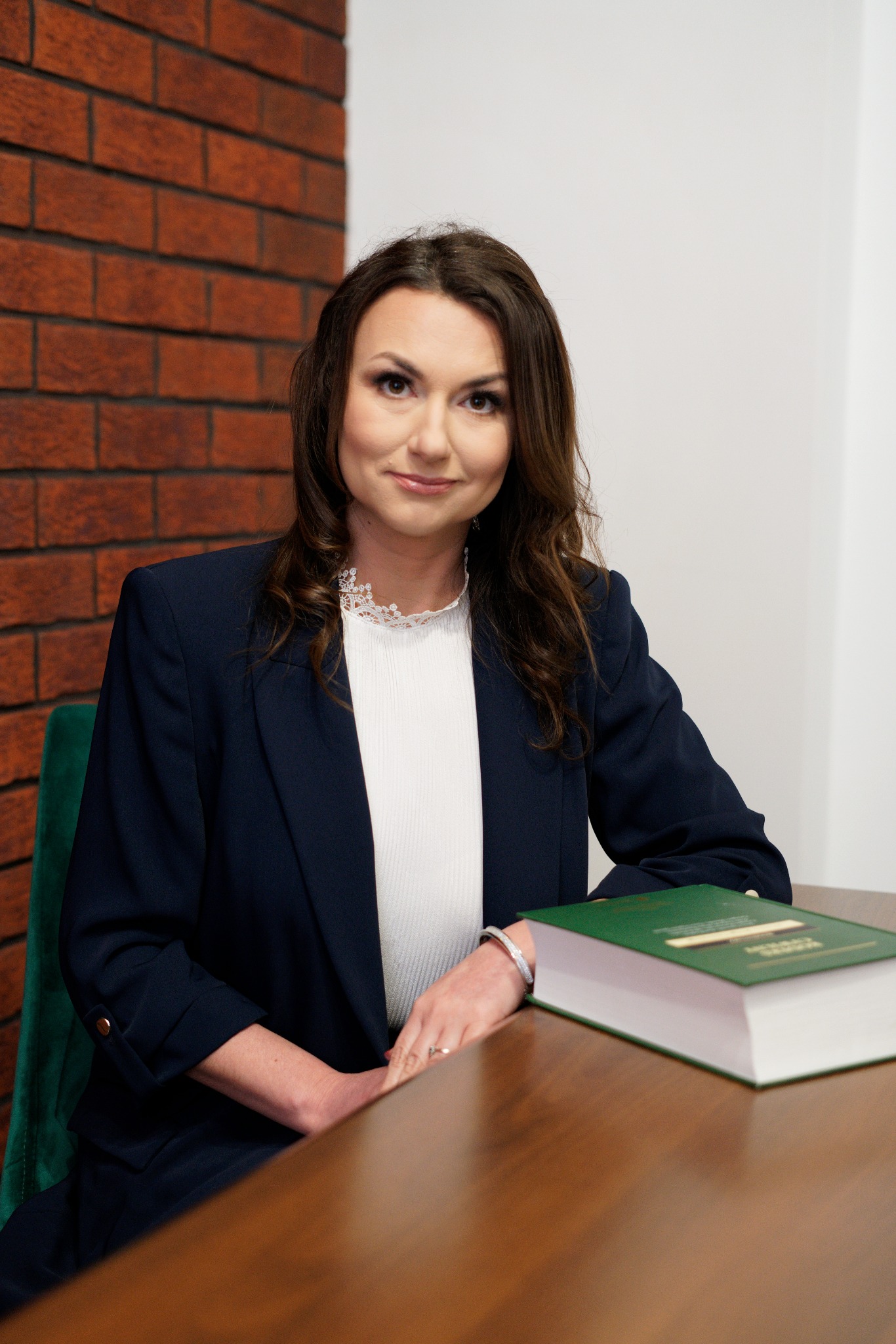 Portret kobiety w granatowej marynarce siedzącej za drewnianym biurkiem z zieloną książką, w tle ceglana ściana