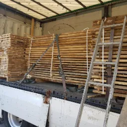 Tarcica ułożona w paczki i zabezpieczona pasami transportowymi w przestrzeni ładunkowej samochodu ciężarowego. Widoczna metalowa drabina oparta o ładunek.