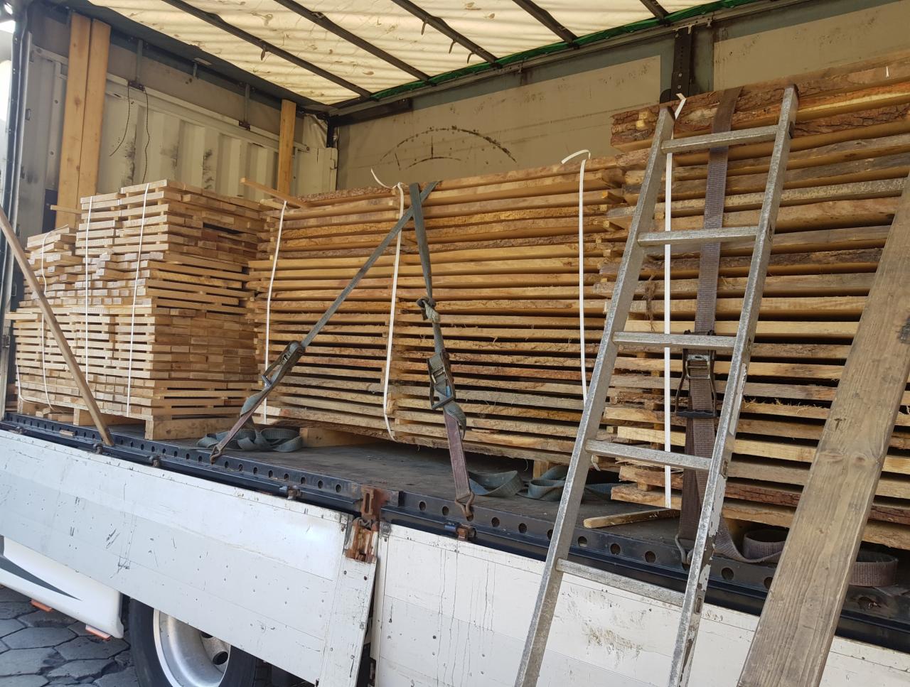 Tarcica ułożona w paczki i zabezpieczona pasami transportowymi w przestrzeni ładunkowej samochodu ciężarowego. Widoczna metalowa drabina oparta o ładunek.