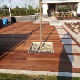 Wykończony taras z drewna egzotycznego z wbudowanym miejscem na drzewo i ścieżką z płyt betonowych, w tle zadaszona przestrzeń wypoczynkowa.