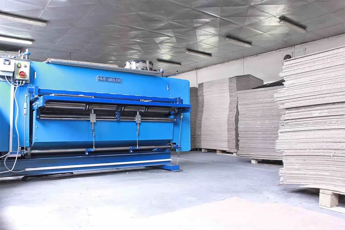 Wnętrze hali produkcyjnej z niebieską maszyną HMB Mikołów do produkcji opakowań kartonowych i ułożonymi na paletach stosami arkuszy tektury.