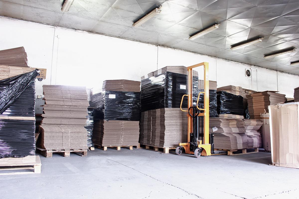 Stosy płaskich, tekturowych arkuszy na paletach, niektóre owinięte czarną folią, z żółtym wózkiem widłowym w tle, w magazynie z metalowym sufitem.