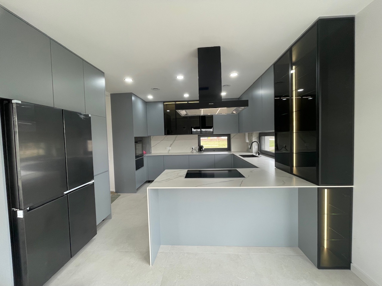 Nowoczesna kuchnia z szarymi i czarnymi szafkami, wyspą z białym blatem i wbudowaną płytą indukcyjną, oświetleniem LED w szafkach i czarnym okapem.