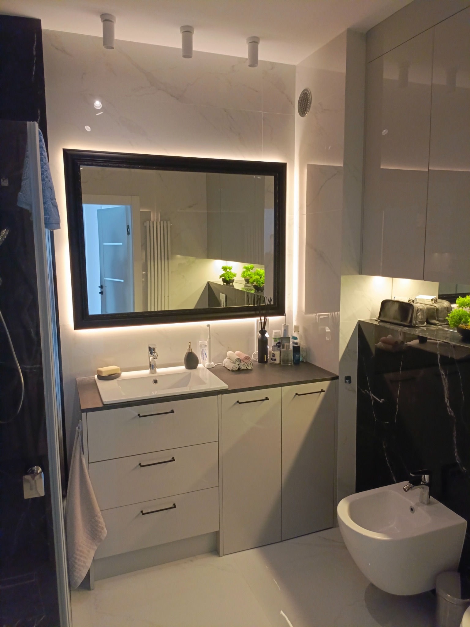 Nowoczesna łazienka z szafką pod umywalkę z szufladami, lustrem z podświetleniem LED, białym bidetem i czarnym marmurem na ścianie.