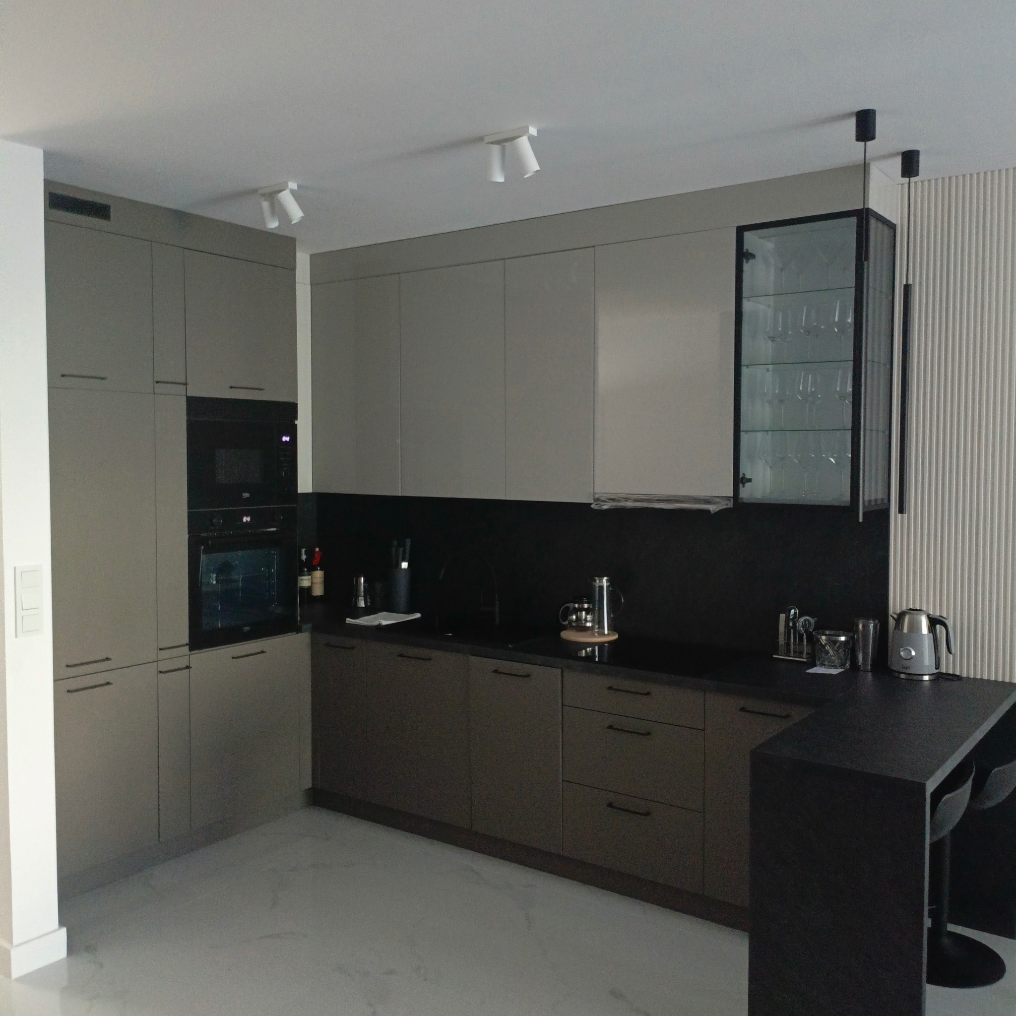 Nowoczesna kuchnia na wymiar w Słupnie z szarymi frontami, czarnym blatem i przeszkloną szafką wiszącą eksponującą kieliszki.