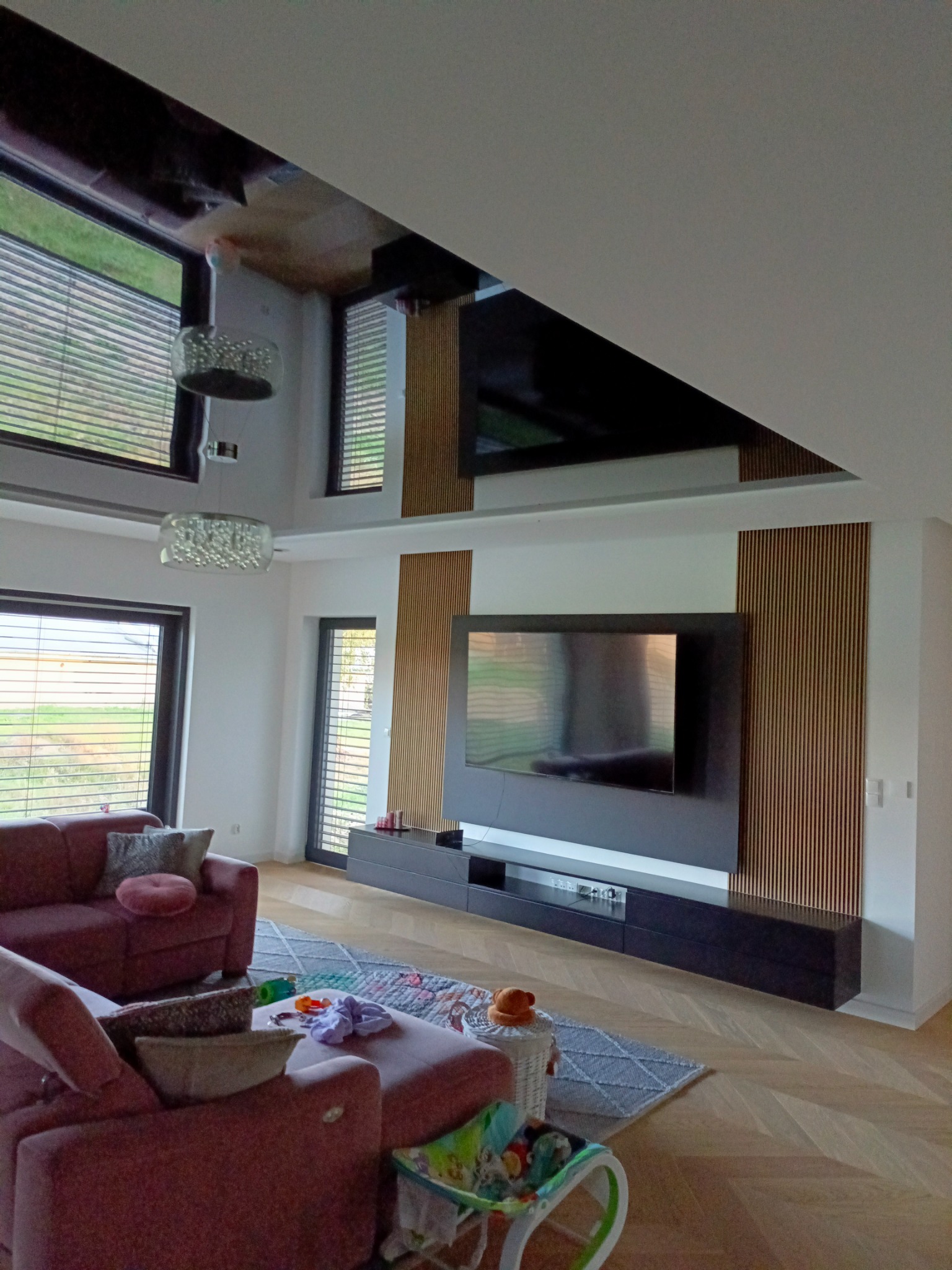 Nowoczesny salon z telewizorem na ścianie, drewniane lamele dekoracyjne, asymetryczny sufit z lustrzanym panelem, kanapa w odcieniach różu i dziecięce zabawki na dywanie.