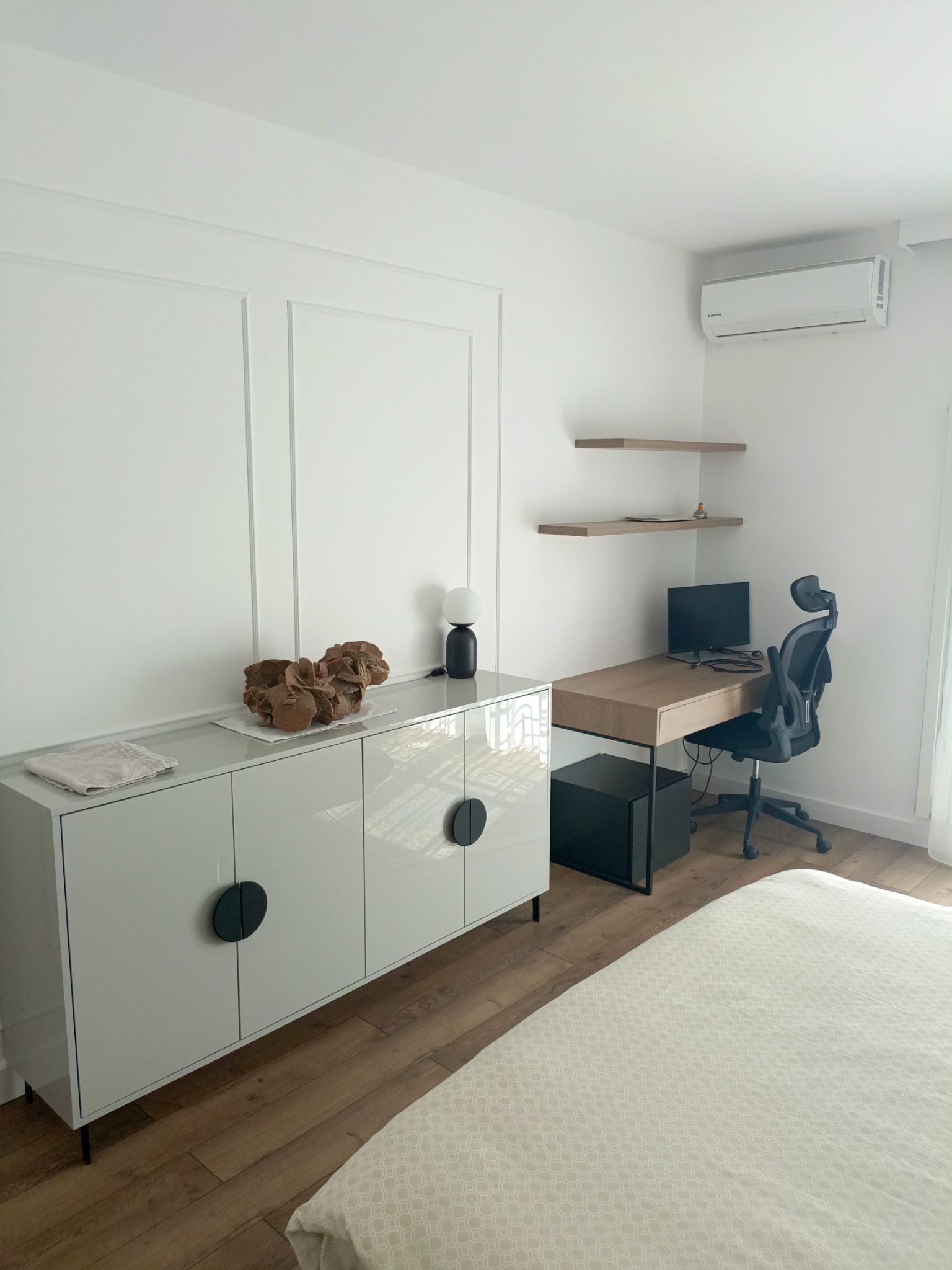 Nowoczesny pokój z białą ścianą z dekoracyjnymi panelami, błyszczącą komodą z okrągłymi uchwytami, biurkiem z komputerem i ergonomicznym krzesłem biurowym, klimatyzator na ścianie.