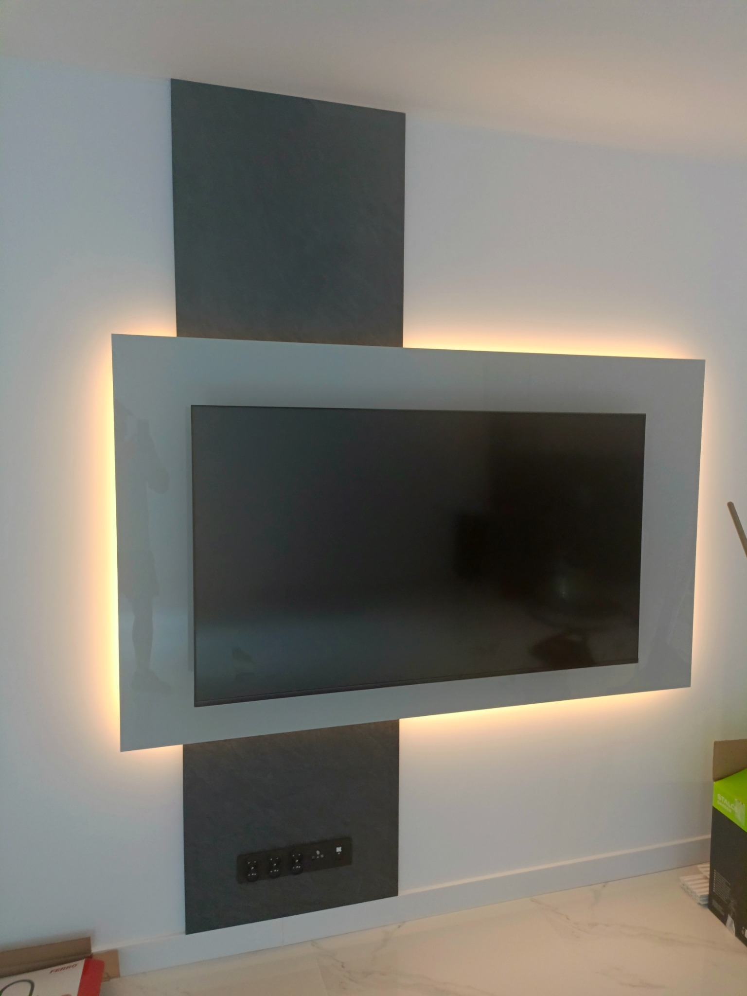 Nowoczesna ściana telewizyjna z podświetleniem LED i panelami dekoracyjnymi w odcieniach szarości, widoczne gniazdka elektryczne.
