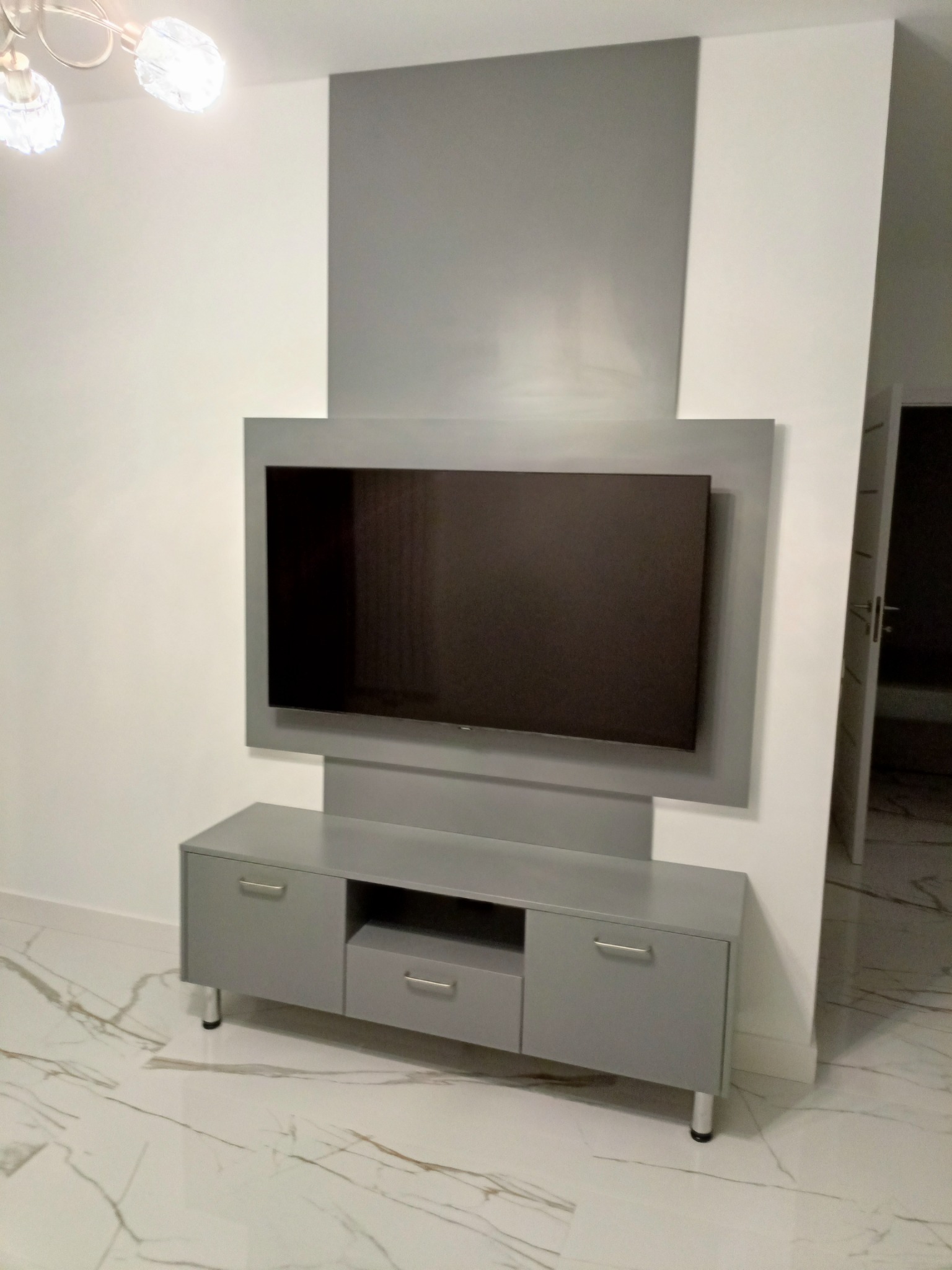 Szara zabudowa ściany z miejscem na telewizor i szafką RTV na metalowych nóżkach, na podłodze marmurkowe płytki.