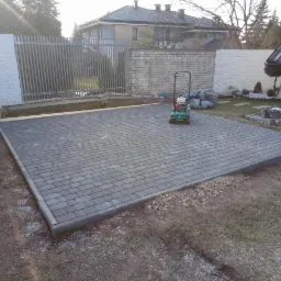 Ułożona szara kostka brukowa na posesji z zagęszczarką w tle, widoczny fragment ogrodu i ogrodzenia.