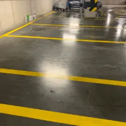 Maszyna czyszcząca posadzkę w garażu podziemnym, widoczne żółte linie wyznaczające miejsca parkingowe, rury pod sufitem i butelki z detergentami przy ścianie.